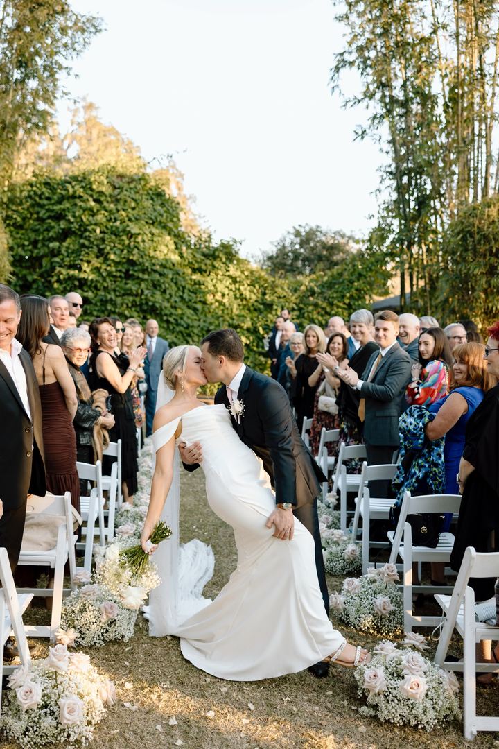The groom dips the bride for a kiss in a garden aisle lined with blush roses and applauding guests