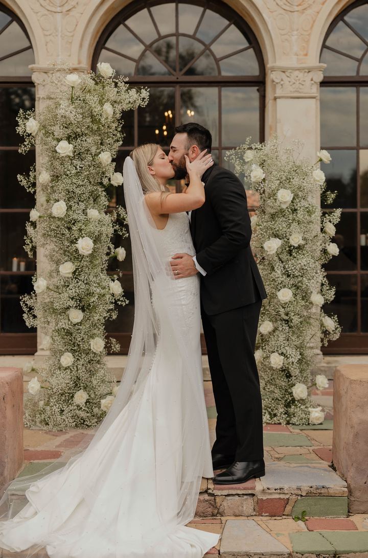A couple's first kiss between columns of white roses and baby's breath, framed by tall arched windows