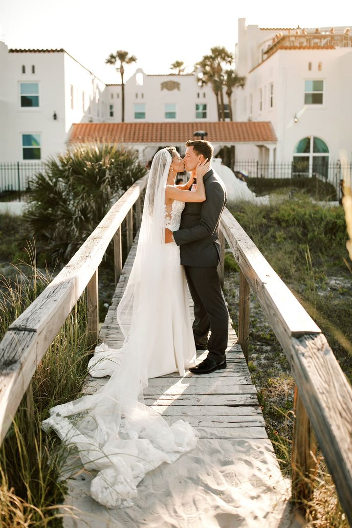 A couple kissing on a beach boardwalk, her cathedral veil trailing across the sand