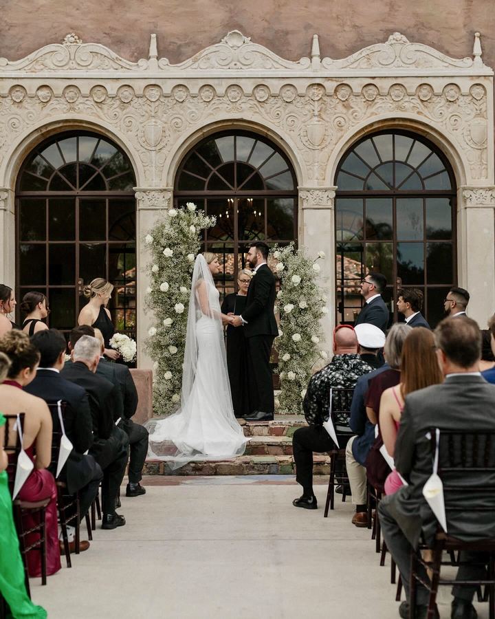 An outdoor ceremony before three carved stone arches, with the officiant standing between the couple as guests look on