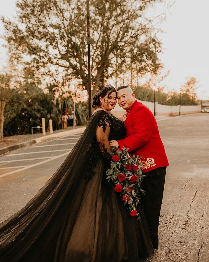 A bride in a black gown and a groom in a red jacket embracing on a quiet road at sunset