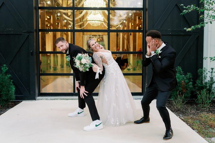 The newlyweds strike a playful pose with a groomsman outside the barn doors