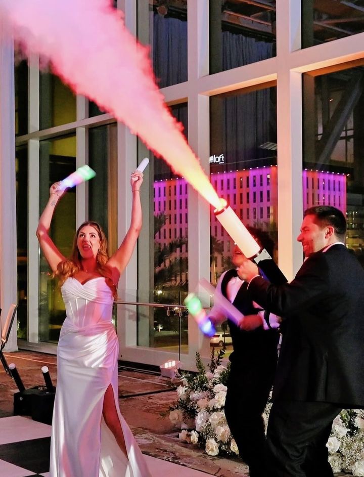 A bride throws her arms up with a light-up baton as a cold spark cannon fires pink smoke over a rooftop dance floor