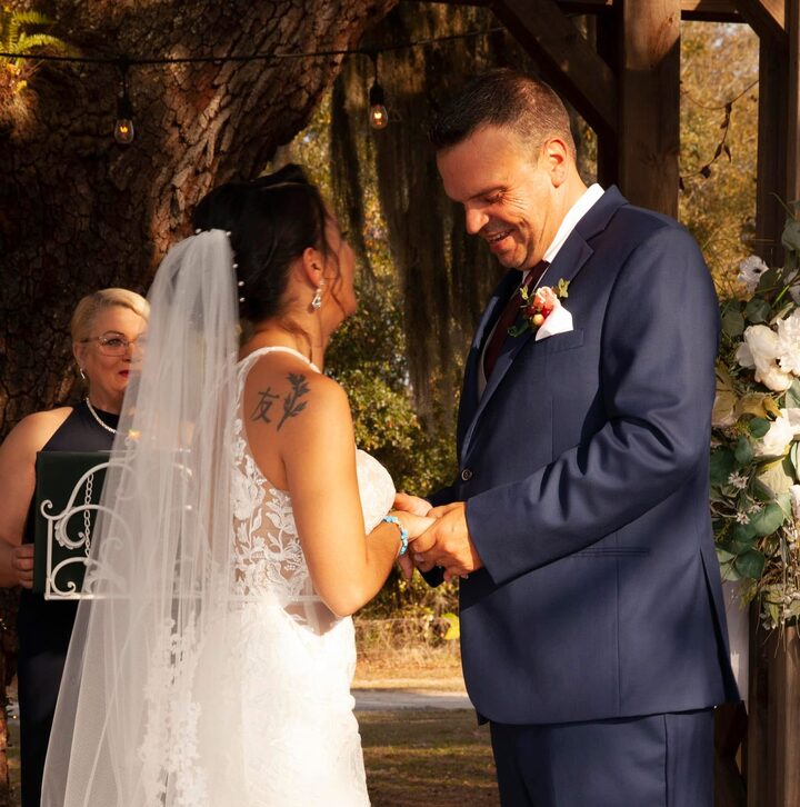Karin looks on as a bride and groom hold hands for their vows beneath a moss-draped live oak hung with string lights