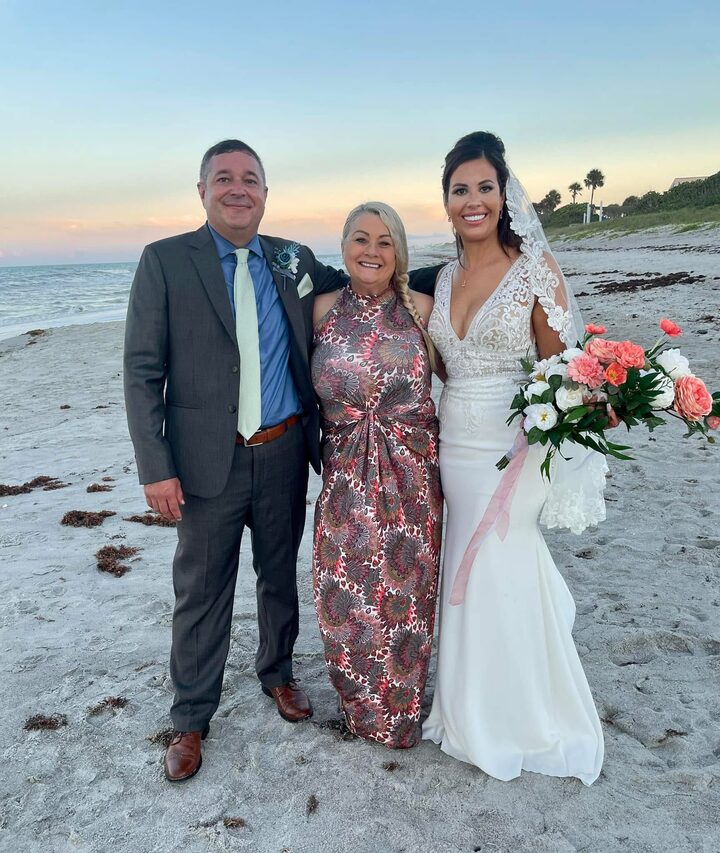 Karin with a bride and groom on the beach at sunset, the bride holding a coral and white bouquet