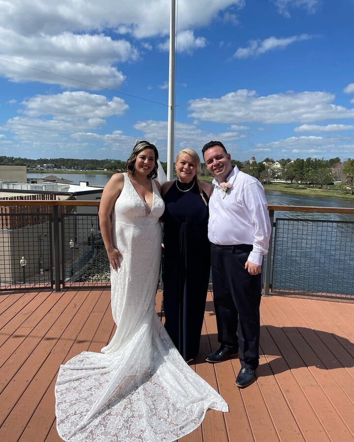 Karin with a bride in a lace gown and a groom on a sunny deck overlooking a lake