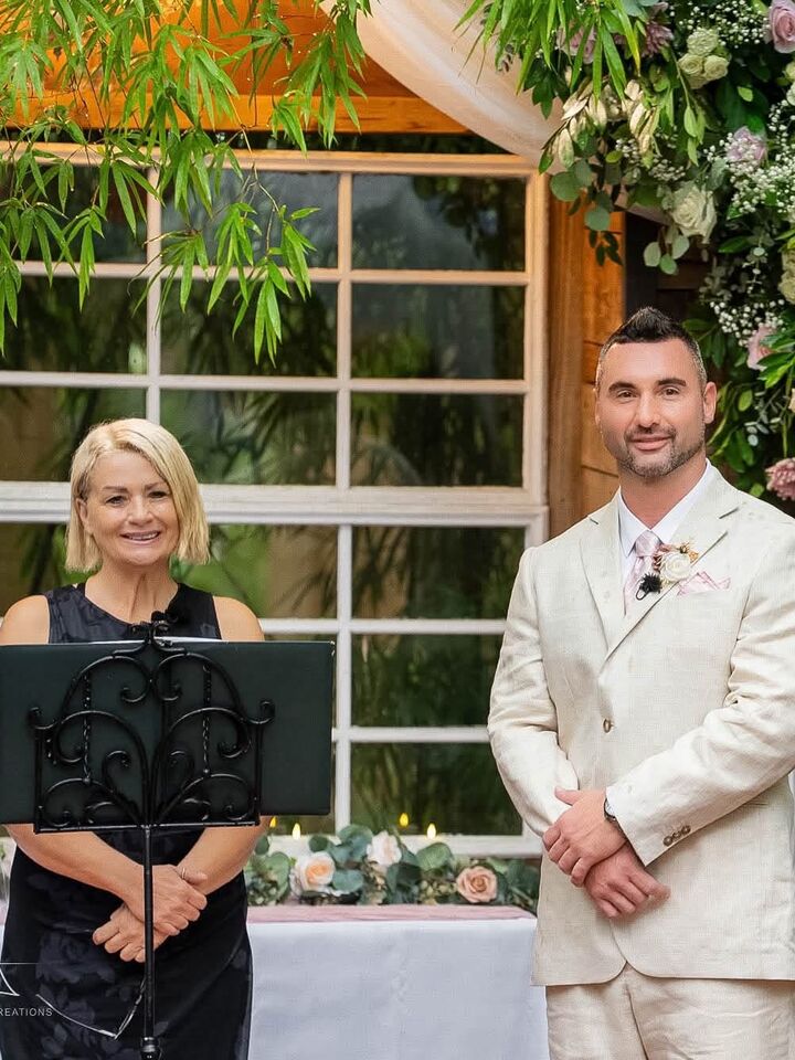 Karin smiling beside a groom in a cream suit at a floral ceremony arch, waiting for his bride
