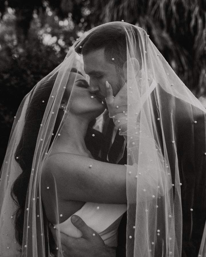 Black and white photo of a couple kissing beneath a pearl-studded veil