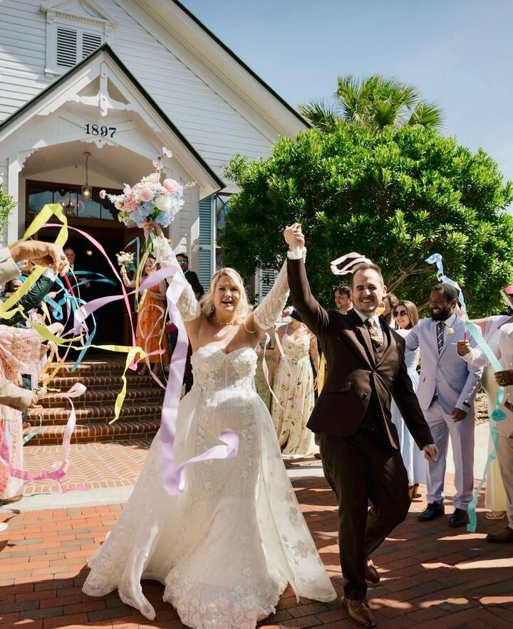 Bride and groom raise their joined hands as guests wave pastel ribbon wands outside a white 1897 chapel