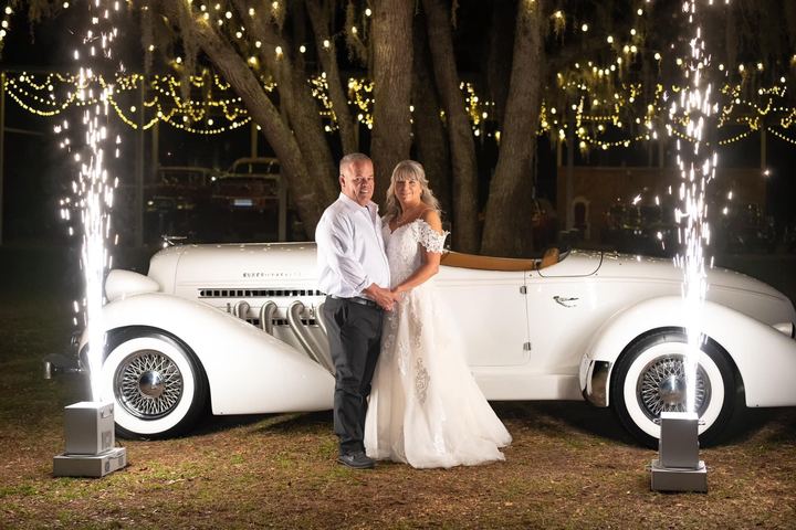 A couple in front of a white vintage roadster, framed by sparkler fountains and string lights
